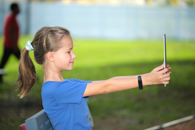 Fröhliches Kindermädchen, das am sonnigen Sommertag auf dem Bildschirm des digitalen Tablets auf der Bank im Park ruht Online-Studienkonzept