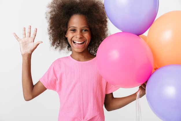 Fröhliches kleines afrikanisches Mädchen mit rosa Bluse steht isoliert über weißer Wand, hat Flosse mit Luftballons, winkende Hand