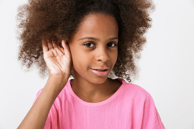 Fröhliches kleines afrikanisches Mädchen mit rosa Bluse steht isoliert über weißer Wand und versucht zu hören
