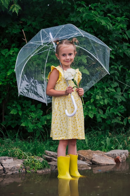 Fröhliches kleines emotionales Mädchen mit transparentem Regenschirm im gelben Kleid und Regenstiefeln im Parksommer