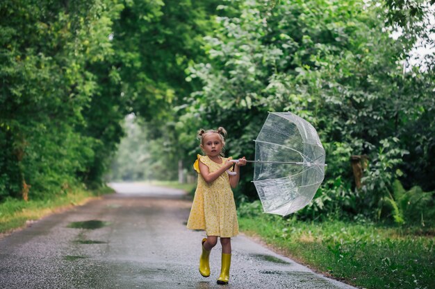 Fröhliches kleines emotionales Mädchen mit transparentem Regenschirm im gelben Kleid und Regenstiefeln im Parksommer