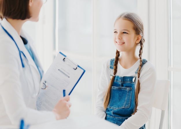 Fröhliches kleines Kind hört aufmerksam auf die Ratschläge und Verschreibungen des Arztes, kommt zum Kinderarzt in die Klinik, hat zwei Zöpfe, trägt einen Jeansoverall, spricht über gesundheitliche Probleme, hat eine Kontrolluntersuchung