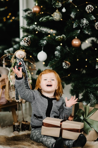 Fröhliches kleines lächelndes Mädchen mit Geschenkbox in der Nähe des Weihnachtsbaums zu Hause Das neue Jahr 2021 Feiertage und Kindheitskonzept