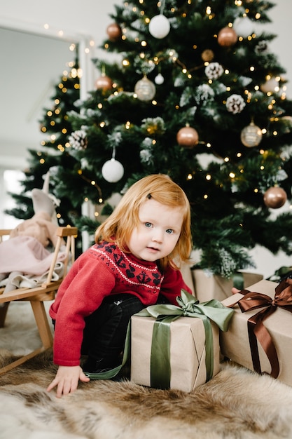 Fröhliches kleines lächelndes Mädchen mit Geschenkbox in der Nähe des Weihnachtsbaums zu Hause Die Feiertage des neuen Jahres 2022