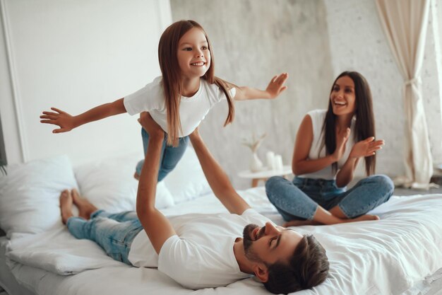 Fröhliches kleines Mädchen breitet ihre Arme weit aus, als würde sie fliegen, während Papa sie hochhebt und Mama zuschaut