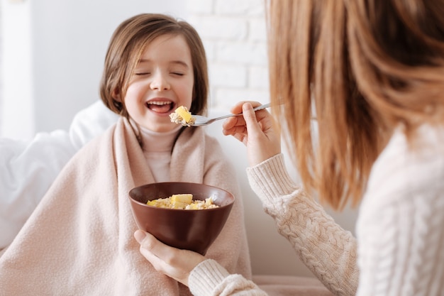 Fröhliches kleines mädchen, das auf der couch sitzt, während ihre liebende mutter ihr frühstück gibt