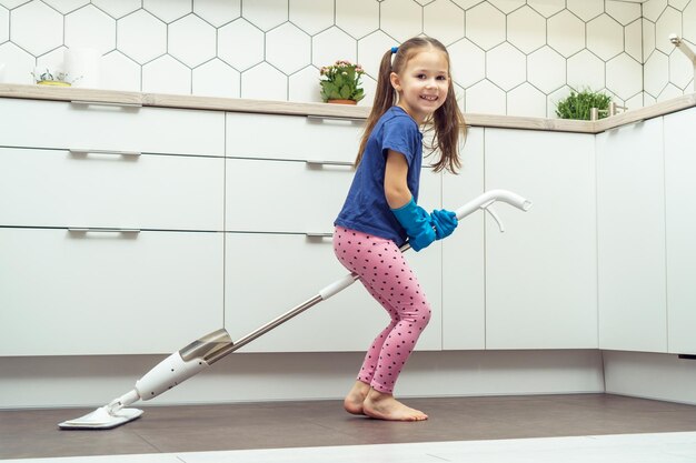 Fröhliches kleines Mädchen, das auf einem modernen Mop sitzt, um den Boden mit einer abnehmbaren Bürste in der Nähe des Küchensets zu waschen. Reinigungskonzept für zu Hause