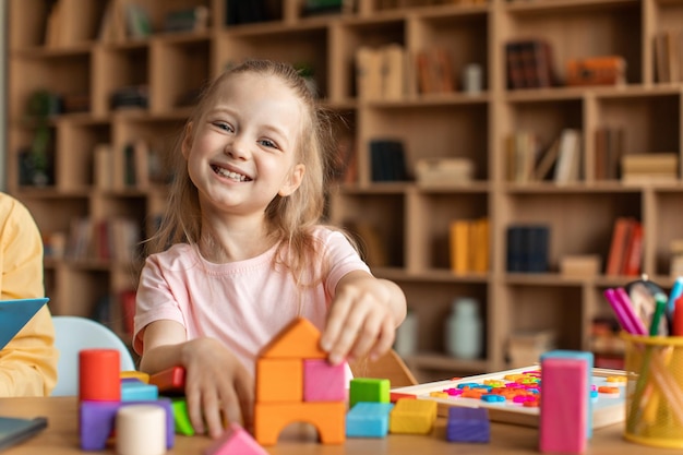 Fröhliches kleines Mädchen, das mit bunten Holzklötzen spielt und in die Kamera lächelt und Tischspiele genießt