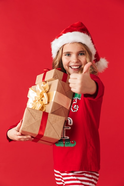 Fröhliches kleines Mädchen, das Weihnachtskostüm trägt, das isoliert steht und Geschenkboxen hält