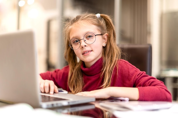 Fröhliches kleines Mädchen, das zu Hause Laptop für Online-Bildung und Heimstudium verwendet und zu Hause mit einem Lächeln in die Kamera schaut. Fernunterricht für Kinder. Soziale Distanz.