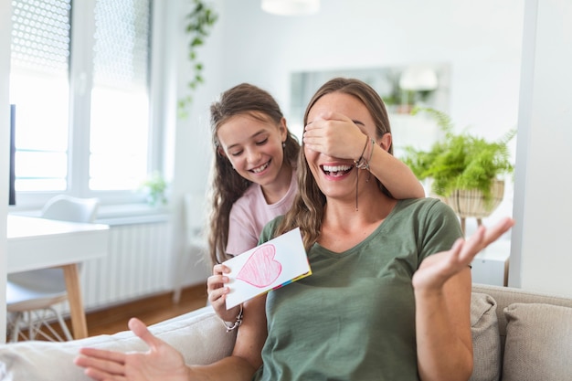 Fröhliches kleines Mädchen gratuliert lächelnder Mutter und gibt Karte mit rotem Herzen während der Feiertagsfeier zu Hause, bedeckt ihre Augen und überrascht sie