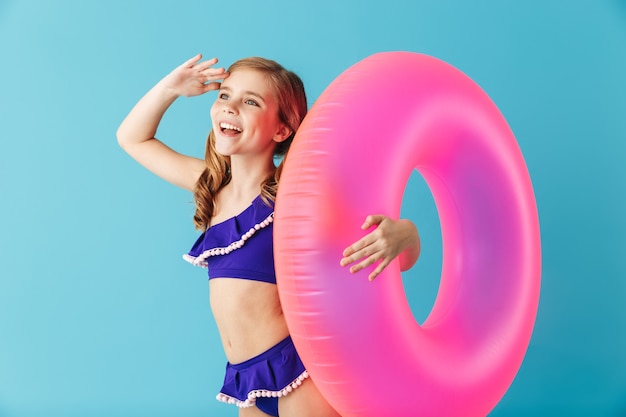 Fröhliches kleines Mädchen im Badeanzug, das isoliert über blauer Wand steht und mit aufblasbarem Ring spielt