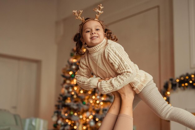 Fröhliches kleines Mädchen in einem Strickpullover hat Spaß zu Hause in der Nähe des Weihnachtsbaums