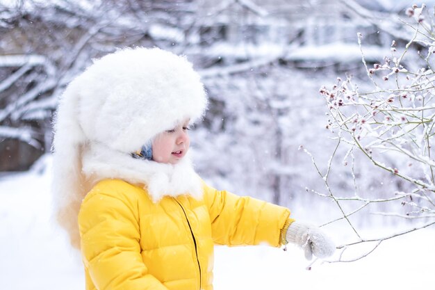 Fröhliches kleines Mädchen in gelber Jacke und flauschigem weißem Hut, das am Wintertag Spaß beim Spielen mit Schnee hat