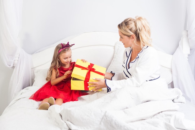 Fröhliches kleines Mädchen in hellem Kleid und Spielzeugkrone auf dem Kopf öffnet Geschenkbox, während sie mit Mutter auf dem Bett sitzt, Frau wünscht Tochter alles Gute zum Geburtstag, grüßt Kind am Feiertagsmorgen, Gratulation