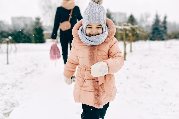 Fröhliches kleines Mädchen lächelt und spielt zusammen mit ihrer Mutter im Park am schneiten Tag. Nettes spielendes Kind fängt mich mit ihrer Mutter im Schnee am Wintertag Weihnachtsstimmung Kindheit