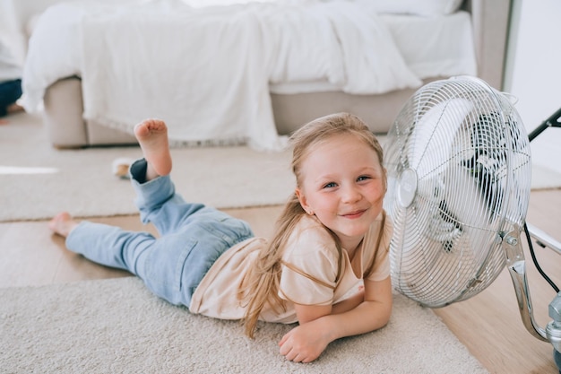 Foto fröhliches kleines mädchen liegt lässig auf dem boden im schlafzimmer in der nähe eines ventilators und entspannt sich an heißen sommertagen