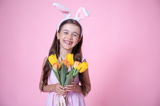 Fröhliches kleines Mädchen mit Osterhasenohren lächelt und hält einen Strauß Tulpen in ihren Händen auf einem rosa Studiohintergrund.