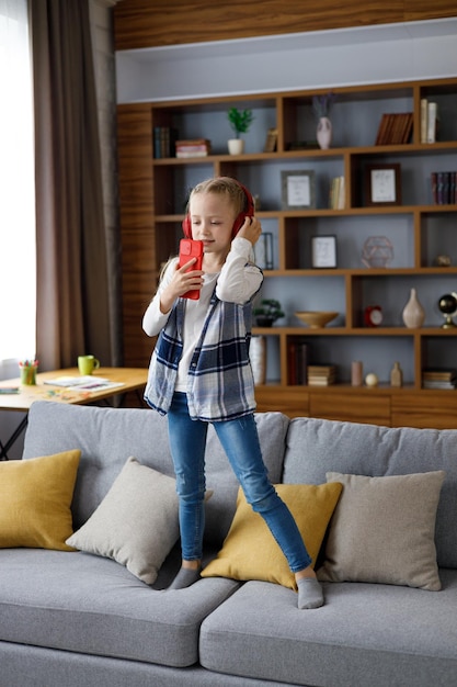 Fröhliches kleines Mädchen mit roten Kopfhörern, das mit einem Smartphone wie einem Mikrofon tanzt und singt Nettes Kind, das Spaß daran hat, auf den Bus zu springen Freizeit glückliches Kind Aufmerksamkeitsdefizit-Hyperaktivitätsstörung
