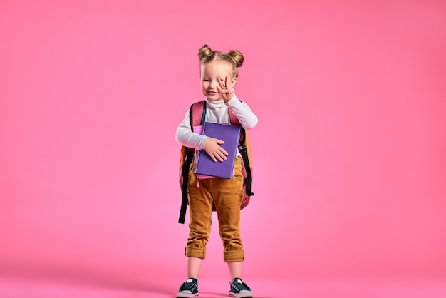 Fröhliches kleines Mädchen mit Rucksack und Büchern, die das Siegeszeichen zeigen, isoliert auf rosafarbenem Hintergrund Kopierbereich