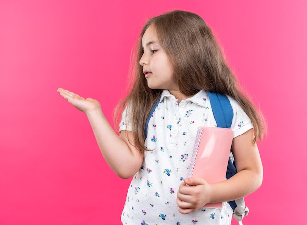 Fröhliches kleines schönes Mädchen mit langen Haaren mit Rucksack, das ein Notizbuch hält und etwas mit dem Arm ihrer Hand präsentiert, das auf rosa steht