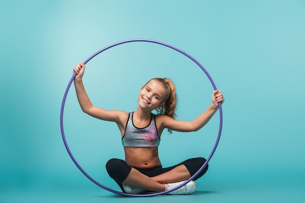 Fröhliches kleines Sportmädchen, das Übungen mit einem Hula Hoop macht, der über blauer Wand lokalisiert wird