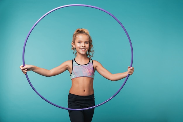 Fröhliches kleines Sportmädchen, das Übungen mit einem Hula Hoop macht, der über blauer Wand lokalisiert wird