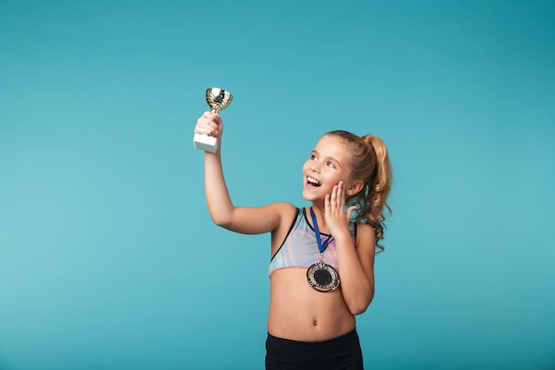 Fröhliches kleines Sportmädchen, das den Sieg feiert, isoliert über der blauen Wand, eine Goldmedaille tragend, eine Trophäe zeigend