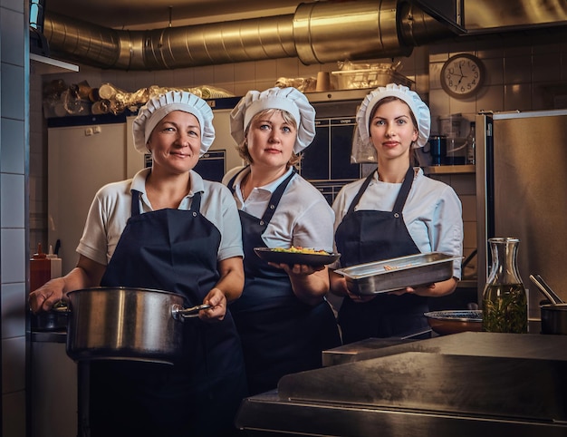 Fröhliches Kochteam in Uniform, das Behälter mit gehacktem Gemüse hält und in der Küche des Restaurants für eine Kamera posiert.
