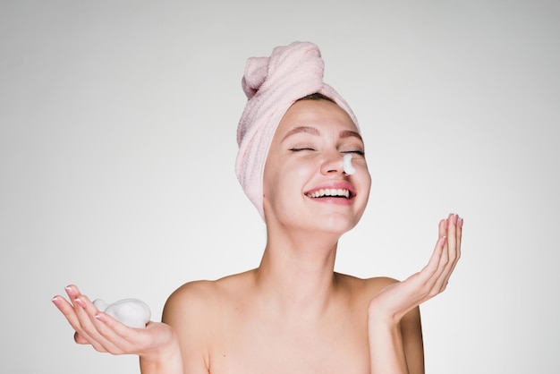 Fröhliches lachendes Mädchen mit rosa Handtuch auf dem Kopf, das Reinigungsschaum auf das Gesicht aufträgt