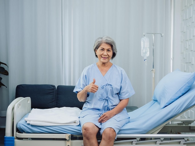 Fröhliches Lächeln Asiatische ältere alte Patientin mit Knieoperation in hellblauem Kleid Daumen hoch, während sie auf dem Bett in der Nähe des medizinischen Infusionsständers mit Kochsalzlösung im Reinraum im Krankenhaus sitzt