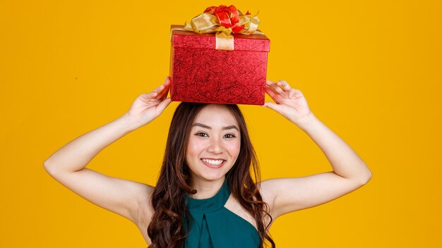 Fröhliches Lächeln Gesicht süßes asiatisches Mädchen mit dunklem Haar, das Geschenkbox über dem Kopf mit entzückender und aufgeregter, Studioaufnahme auf gelbem Hintergrund hält. Feiern und Festivalkonzept.