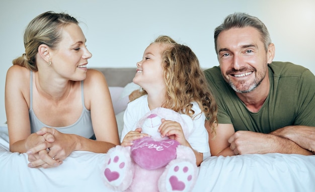 Fröhliches Lächeln und Familie, die sich auf einem Bett entspannen, während sie sich in ihrem ruhigen und komfortablen Schlafzimmer verbinden Glück, Liebe und Fürsorge zwischen Eltern und einem Mädchen, das einen Teddy hält, während es sich in einem modernen Zuhause ausruht