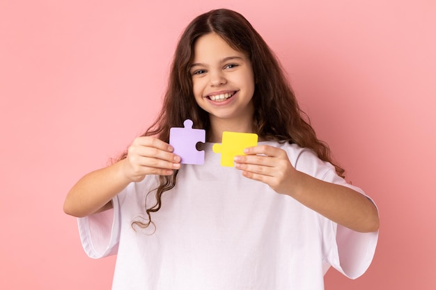 Fröhliches lächelndes kleines Mädchen mit gelben und violetten Puzzleteilen, die Aufgaben lösen und in die Kamera schauen