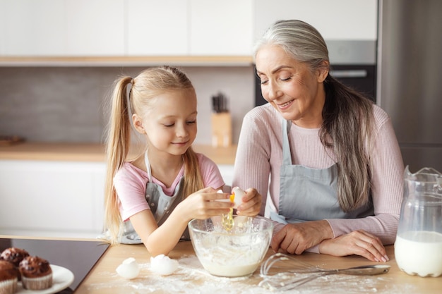 Fröhliches lächelndes kleines Mädchen und ältere Frau in Schürzen machen Teigbruchei im minimalistischen Kücheninterieur