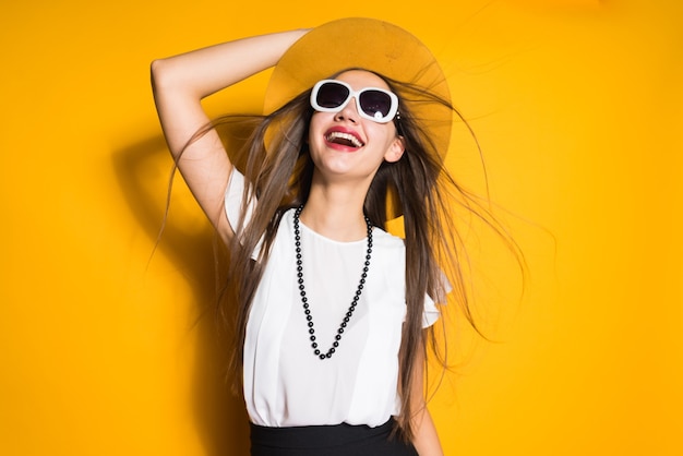 Fröhliches lächelndes Mädchen mit langen Haaren posiert, in trendigem Hut und Sonnenbrille