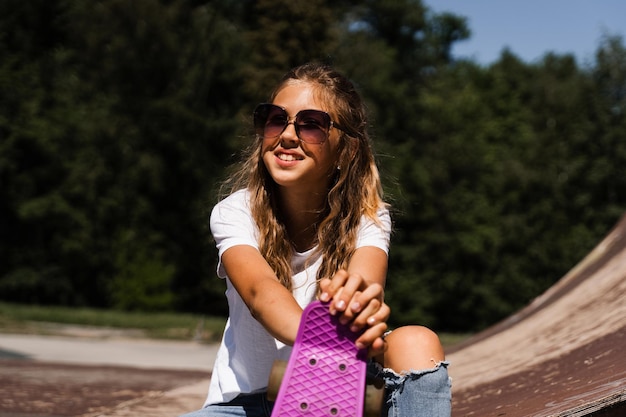 Fröhliches lächelndes Mädchen mit Skateboard, das auf dem Skate-Spielplatz sitzt und Spaß hat Extremsport-Lifestyle Lachendes Kind mit Skateboard, das auf der Sportrampe posiert