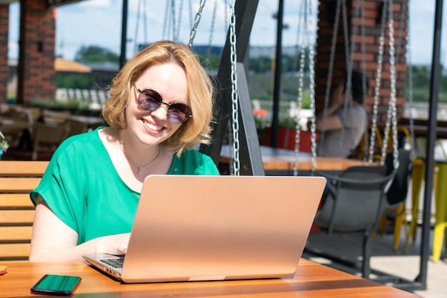 Fröhliches lächelndes Mädchen mit Sonnenbrille, das eine Anwendung auf einem digitalen Laptop in einem Straßencafé verwendet