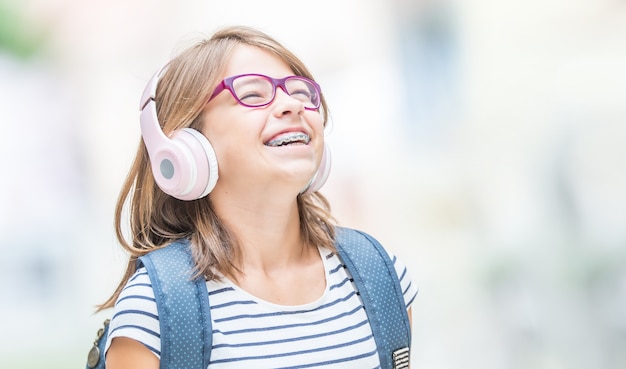 Fröhliches lächelndes Schulmädchen mit Zahnspangen und Brille, die Musik von Kopfhörern hört. Kieferorthopäde und Zahnarztkonzept.