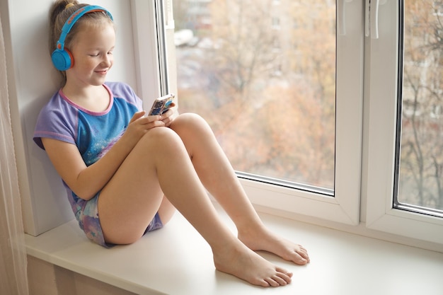 Fröhliches lächelndes süßes Mädchen mit Smartphone in den Händen sitzt auf der Fensterbank und hört Musik über Kopfhörer