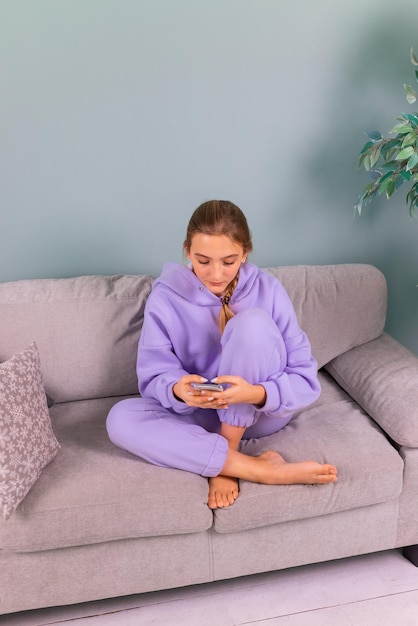 Fröhliches lächelndes Teenager-Mädchen mit ihrem Smartphone, das soziale Netzwerke durchsucht und entspannt auf einem Sofa zu Hause sitzt Online-Bildungsstudie von zu Hause aus