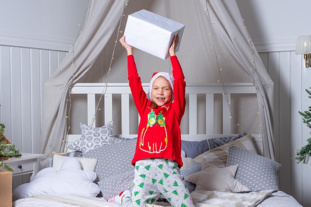 Fröhliches, lustiges Babymädchen in Schlafanzug und Weihnachtsmütze wirft und fängt am Weihnachtsmorgen eine Geschenkbox im Bett