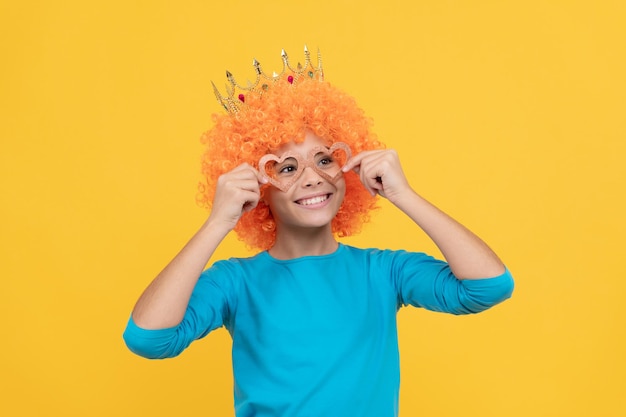 Fröhliches, lustiges Kind mit lockiger Perücke und Krone stellt sich vor, eine Königin zu sein. Fröhliches Kind trägt ein Diadem. Teenager-Mädchen mit Partybrille ist ein Clown