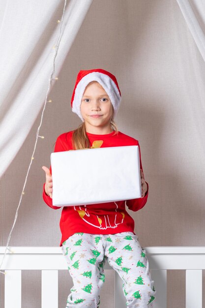 Fröhliches, lustiges Mädchen im Schlafanzug und Weihnachtsmütze, das am Weihnachtsmorgen eine Geschenkbox im Bett hält