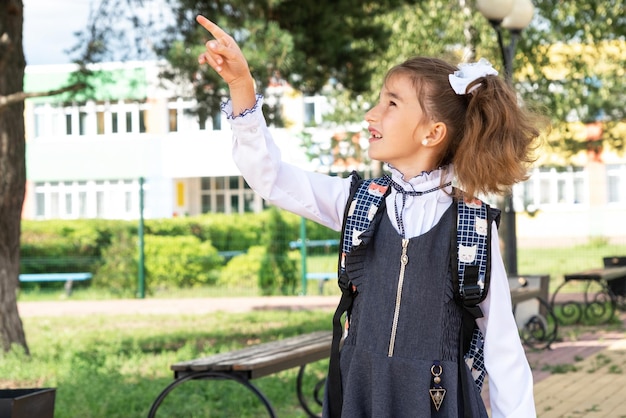 Foto fröhliches, lustiges mädchen in schuluniform mit weißen schleifen auf dem schulhof zeigt mit dem finger zurück zur schule 1. september glückliche schülerin mit rucksack grundschulbildung grundklasse