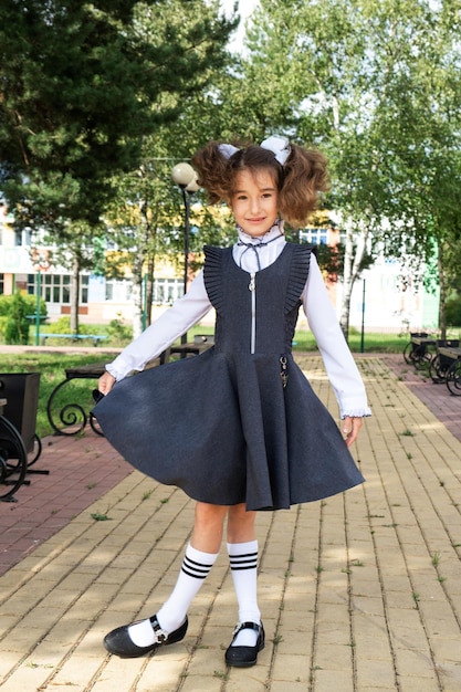 Fröhliches, lustiges Mädchen mit zahnlosem Lächeln in Schuluniform mit weißen Schleifen auf dem Schulhof. Zurück zur Schule, 1. September. Ein glücklicher Schüler. Grundschulbildung, Grundschule. Porträt eines Schülers