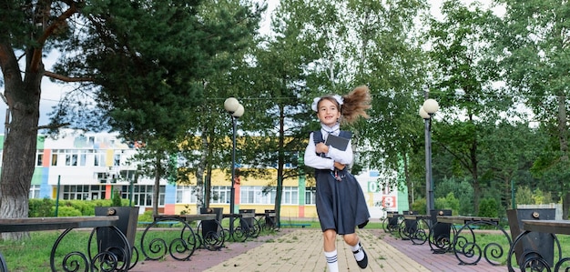 Fröhliches, lustiges Mädchen mit zahnlosem Lächeln in Schuluniform mit weißen Schleifen, das auf dem Schulhof rennt. Zurück zur Schule, 1. September. Glückliche Schülerin mit Rucksack. Bewegung in der Grundschule