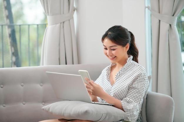 Fröhliches Mädchen, das bequem auf beigem Sofa sitzt und Laptop-SMS online verwendet Kopierbereich