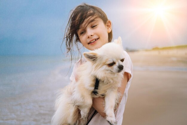 Fröhliches Mädchen, das ihren kleinen Hund umarmt, züchtet Chihuahua an der Küste vor blauem Himmel, Nahaufnahme der besten Freunde