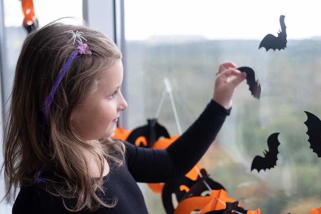 Fröhliches Mädchen im Hexenkostüm, das Fenster im Zimmer mit schwarzen Fledermäusen für Halloween dekoriert